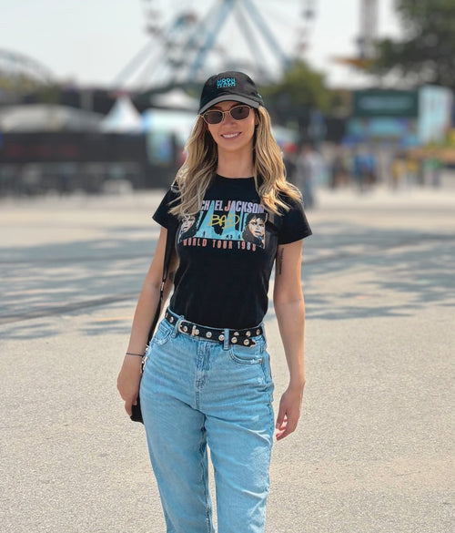 A woman walking outdoors wearing a black Michael Jackson 'World Tour 1985' graphic t-shirt, light blue jeans, a black studded belt, and a black baseball cap.