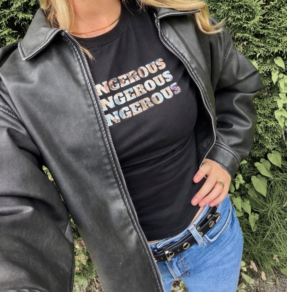 A person wearing a black faux-leather zip-up jacket over a black graphic T-shirt with the word 'DANGEROUS' repeated in a rainbow gradient, paired with blue jeans, standing outdoors in front of green foliage.