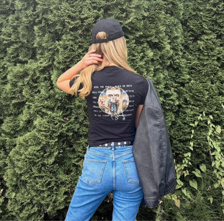 A woman stands with her back to the camera in front of a green hedge, wearing a black graphic t-shirt, blue jeans, a black baseball cap, and carrying a black leather jacket over her shoulder.