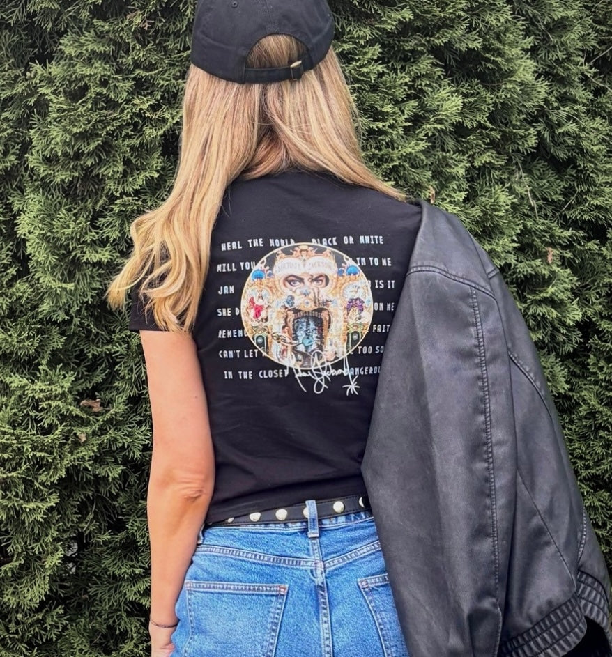 A woman viewed from behind wearing a black graphic t-shirt with a detailed multicolor mandala print and text, a black baseball cap, and a black leather jacket draped over her shoulder, set against a green hedge background.