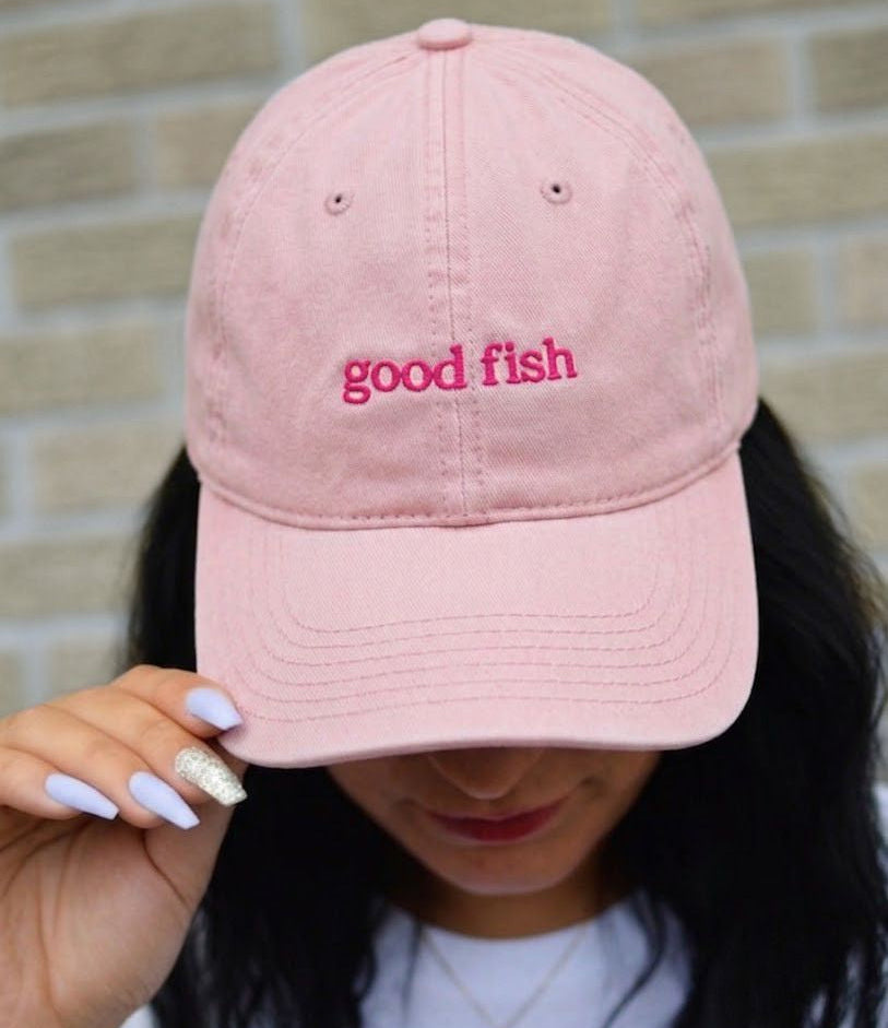 A person wearing a light pink baseball cap with 'good fish' embroidered in pink on the front, holding the brim with one hand against a light brick wall background.
