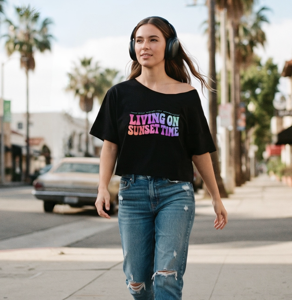 A woman walking on a palm-tree-lined street wearing a black off-the-shoulder graphic T-shirt with the text 'LIVING ON SUNSET TIME' in pink, purple, and blue, paired with distressed blue jeans and black headphones.