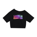 A woman walking on a palm-tree-lined street wearing a black off-the-shoulder graphic T-shirt with the text 'LIVING ON SUNSET TIME' in pink, purple, and blue, paired with distressed blue jeans and black headphones.