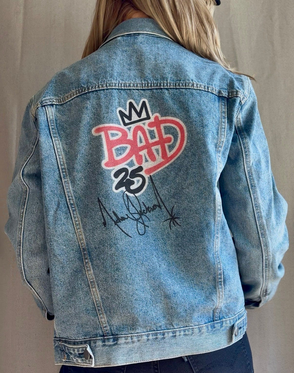 A person viewed from behind wearing a light-wash blue denim jacket with a large back graphic that reads 'BAD 35' in red and black, along with a black signature-style print. The photo is cropped from the shoulders to the hips against a plain light neutral backdrop.