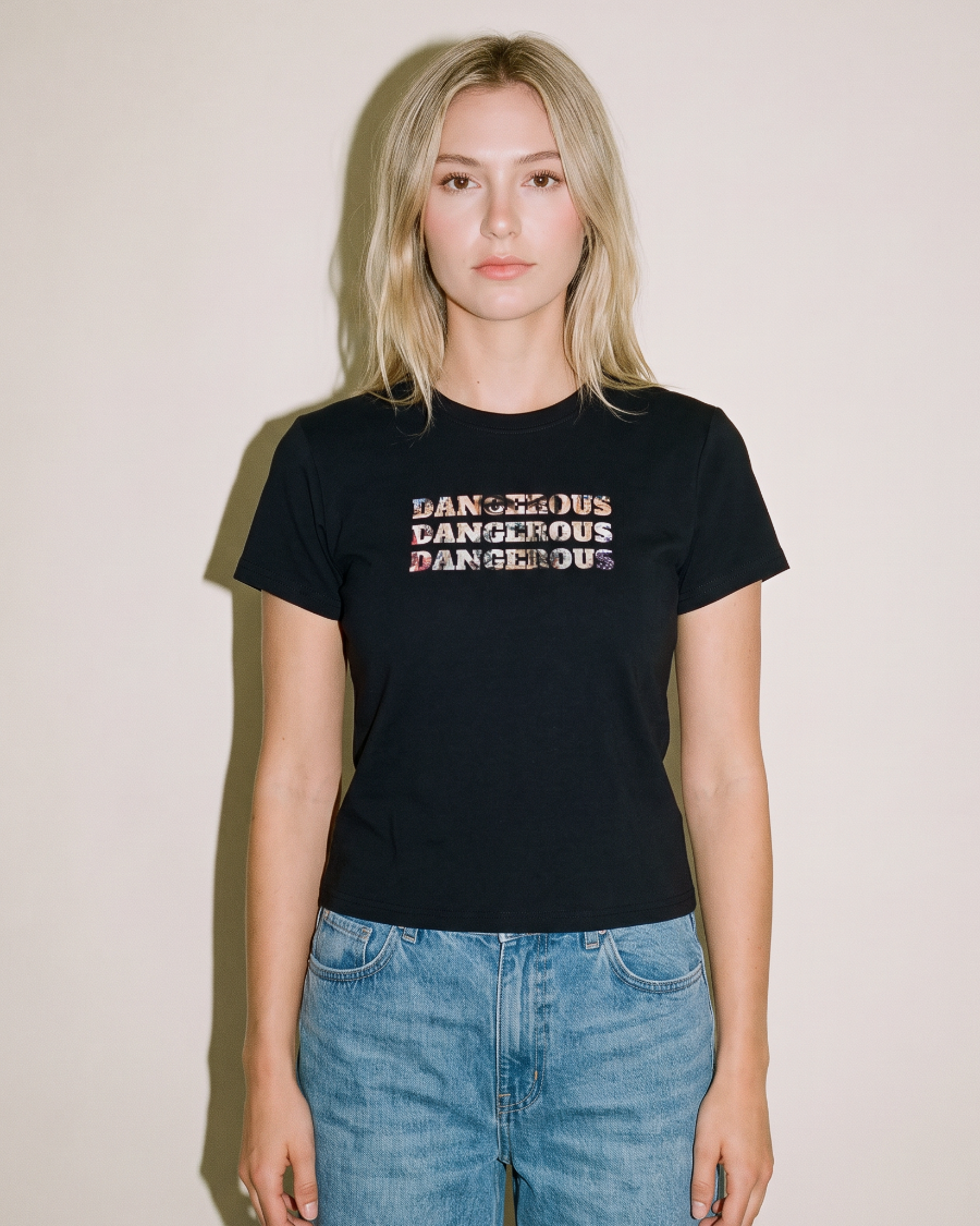 A blonde woman wearing a black short-sleeve graphic t-shirt with the word 'DANGEROUS' repeated in a distressed white and pink font, paired with light blue jeans, standing against a plain beige wall.