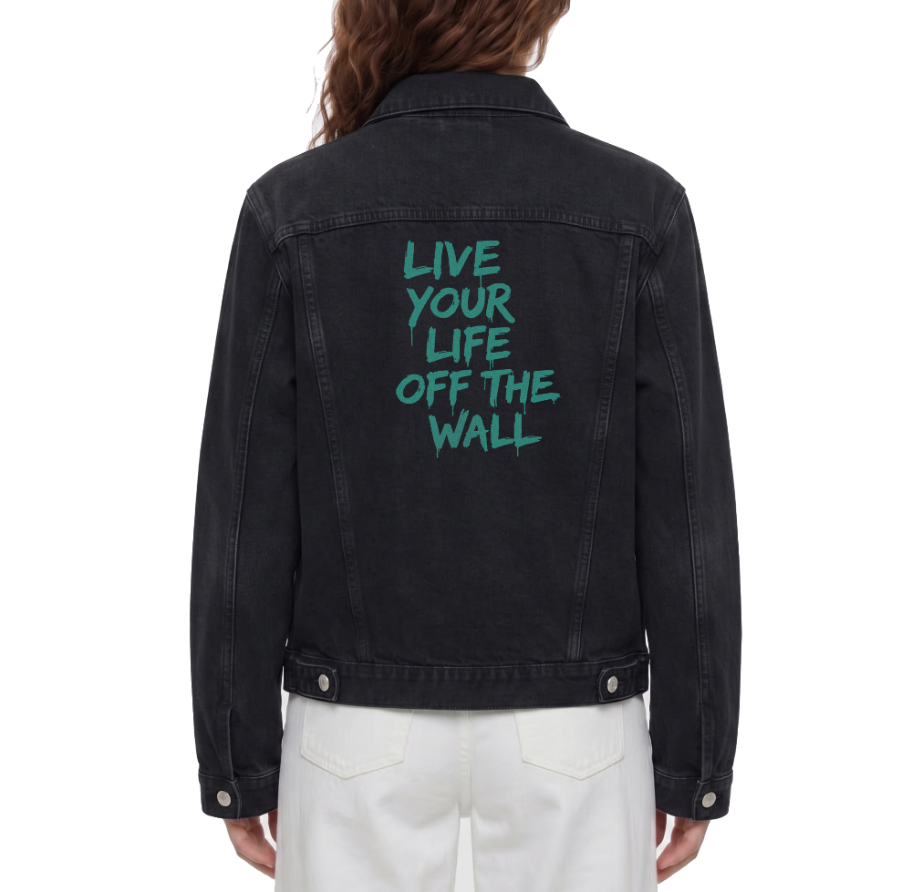 A person shown from the back wearing a washed black denim jacket with the phrase 'LIVE YOUR LIFE OFF THE WALL' printed in teal across the upper back, paired with white pants against a plain white studio background.